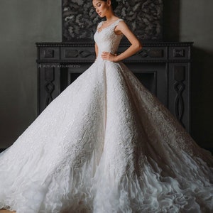 May include: A white wedding dress with a fitted bodice and a full skirt. The skirt has a layer of ruffles at the bottom. The dress is decorated with intricate lace patterns.