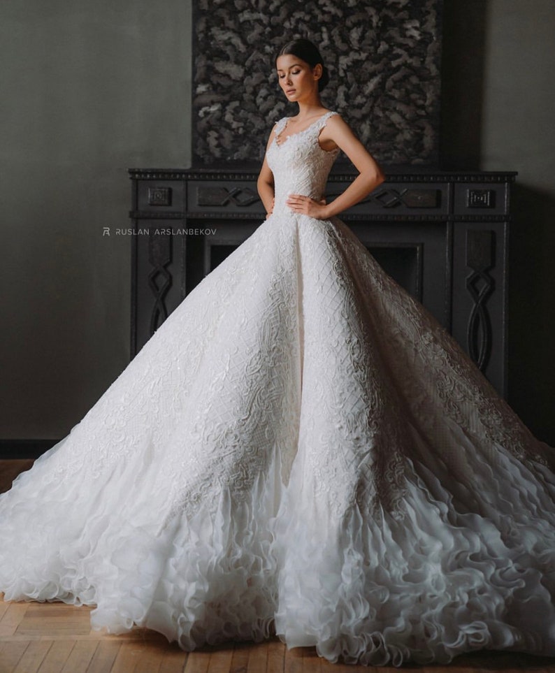 May include: A white wedding dress with a fitted bodice and a full, flowing skirt. The skirt has a layer of ruffles at the bottom. The dress is decorated with intricate lace and beading.