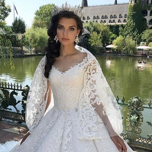 May include: A white lace wedding dress with a v-neckline and a long train. The dress has a fitted bodice and a full skirt. The bride is wearing a tiara and earrings.
