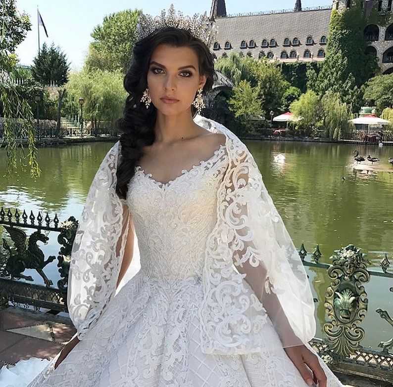 May include: A white lace wedding dress with a v-neckline and a long train. The dress has a fitted bodice and a full skirt. The bride is wearing a tiara and earrings.