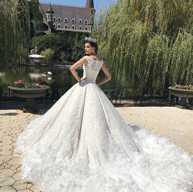 May include: A white wedding dress with a lace bodice and a full skirt. The dress has a long train and is decorated with ruffles. The dress is being worn by a woman in a garden setting.