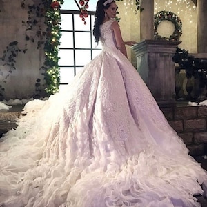 May include: A white wedding dress with a lace bodice and a full skirt. The skirt is made of layers of tulle and has a ruffled hem. The dress is being worn by a woman with long dark hair and a tiara.
