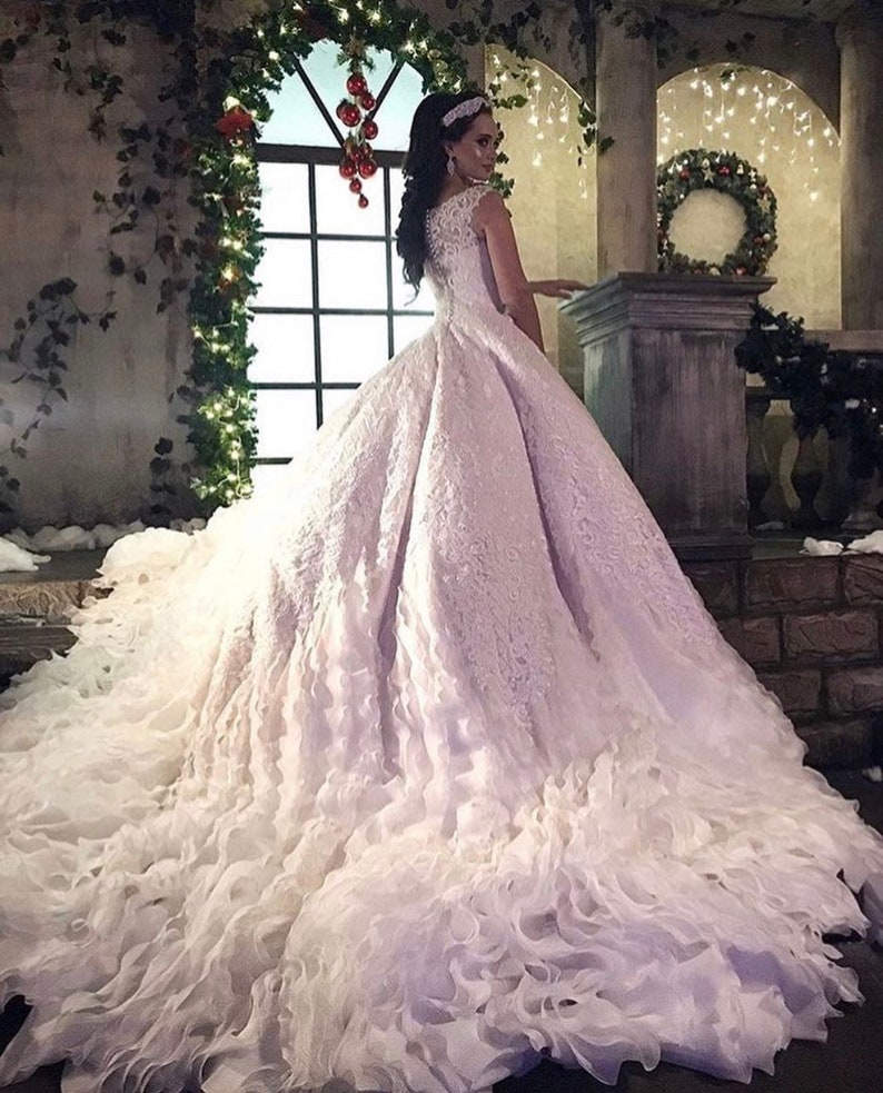 May include: A white wedding dress with a lace bodice and a full skirt. The skirt is made of layers of tulle and has a ruffled hem. The dress is being worn by a woman with long dark hair and a tiara.