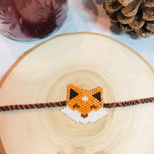 May include: A brown and white beaded fox charm bracelet with a brown cord. The bracelet is on a wooden surface.
