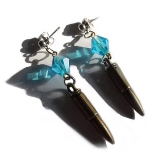 May include: A pair of dangle earrings featuring a bullet design. Each earring has a silver-tone stud, a light blue crystal, and a bronze-colored bullet pendant. The earrings are set against a white background.