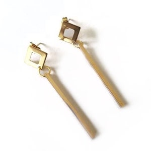 May include: A pair of gold-colored earrings. Each earring features a square top with a cutout and a long, rectangular dangle. The earrings are displayed on a white background.