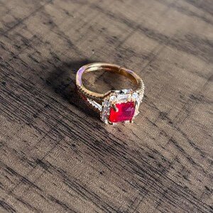 May include: A gold ring featuring a large red square-shaped gemstone surrounded by smaller clear gemstones.