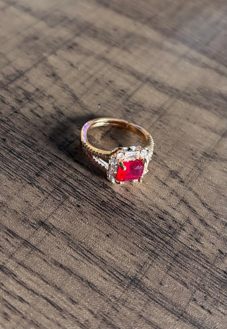 May include: A gold ring featuring a large red square-shaped gemstone surrounded by smaller clear gemstones.