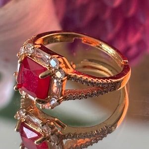 May include: A gold ring with a large, square, red gemstone surrounded by smaller clear gemstones. The ring is set on a white surface.