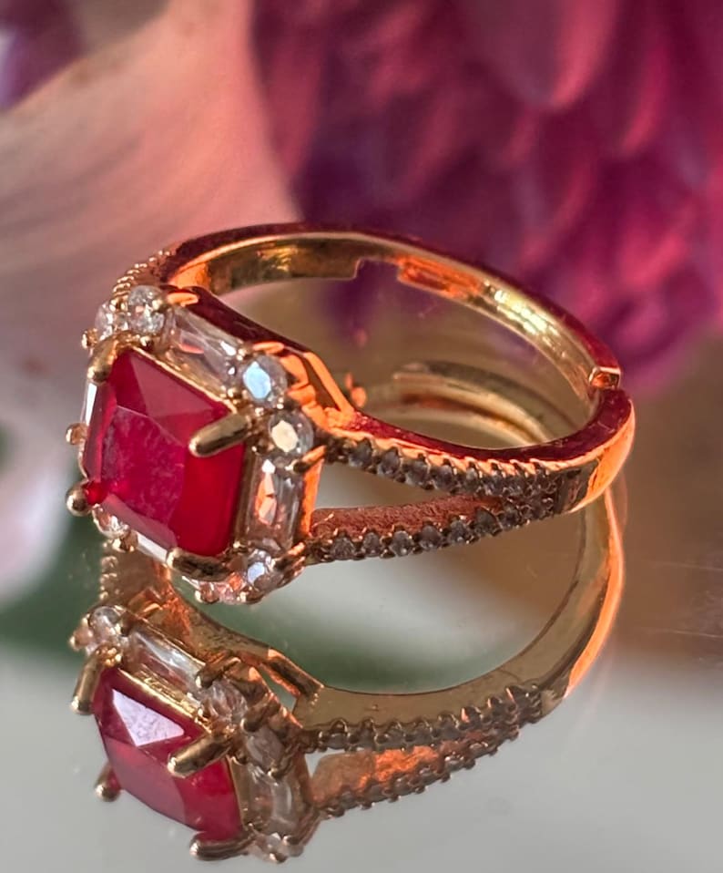 May include: A gold ring featuring a large, square, red gemstone surrounded by smaller clear gemstones. The ring is displayed on a white surface.