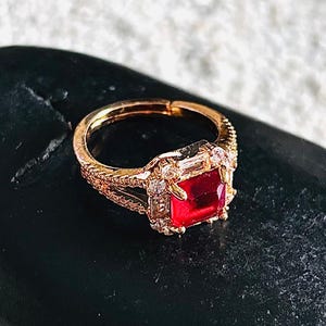 May include: Gold ring featuring a square red gemstone surrounded by smaller clear gemstones.