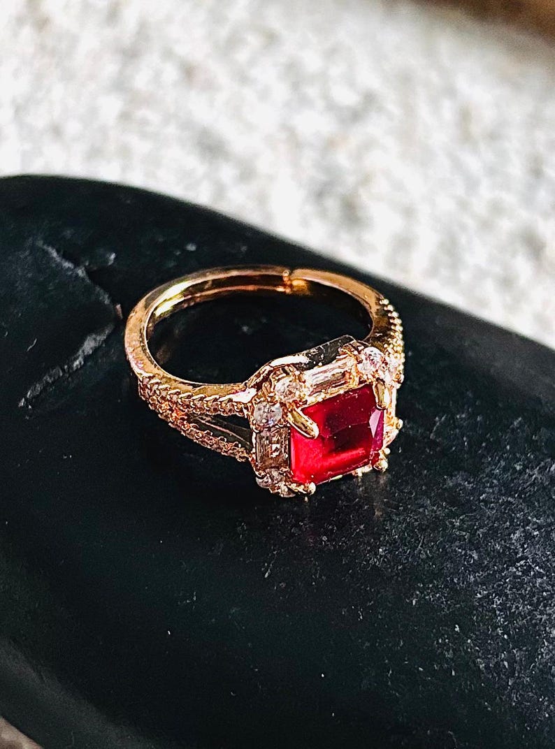 May include: Gold ring featuring a square red gemstone surrounded by smaller clear gemstones.