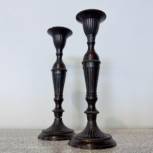 Könnte beinhalten: Zwei antike, dunkelbraune Metallkerzenständer. Die Kerzenhalter haben ein geriffeltes Design an den Stielen und einen dekorativen Sockel. Die Kerzenhalter sind unterschiedlich hoch, einer ist größer als der andere. Der Hintergrund ist eine schlichte, weiße Wand.
