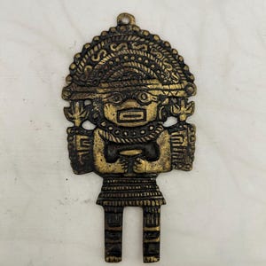 May include: A bronze-colored pendant in the shape of a stylized figure. The pendant features intricate details, including a headdress, facial features, and a patterned skirt. The figure's arms are crossed, and it has two rectangular legs. The pendant has a loop at the top for hanging.