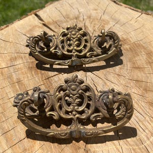 May include: Two antique brass drawer pulls with ornate floral and scroll designs. Each pull features a central decorative element flanked by two smaller, curved handles. The brass has a warm, aged patina, suggesting vintage craftsmanship.