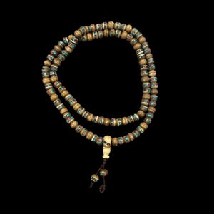 Puede incluir: Un collar de cuentas sobre un fondo oscuro. El collar presenta múltiples hilos de cuentas redondas de color dorado, intercaladas con detalles turquesa y rojos. Un colgante y borla blancos, con forma de hueso, cuelgan del collar.