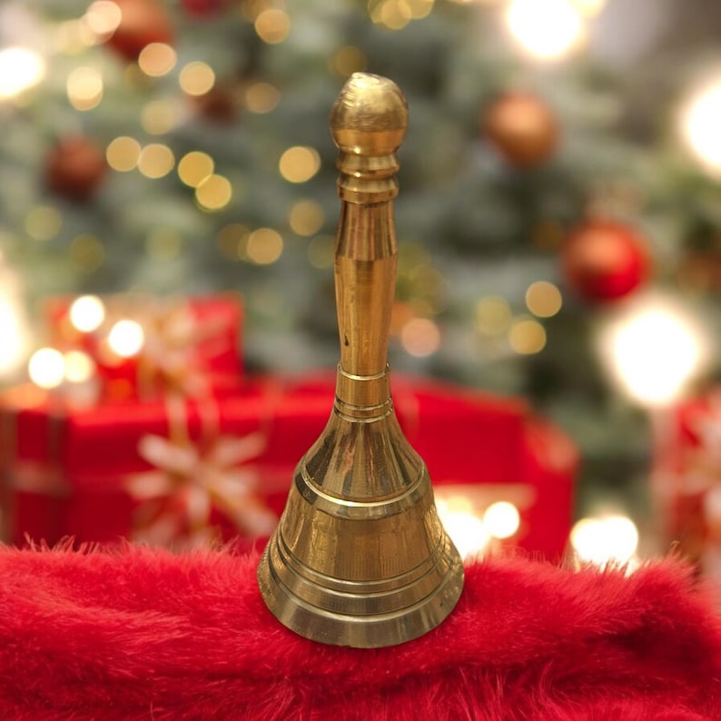 Pode incluir: Um sino dourado com o topo arredondado e uma base larga. O sino est&aacute; sentado em uma superf&iacute;cie vermelha e felpuda. O fundo est&aacute; desfocado e mostra uma &aacute;rvore de Natal com luzes e enfeites.