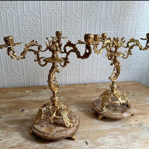 May include: Two antique gold candelabras with multiple arms. Each candelabra features a marble base and elaborate, swirling patterns. The candle holders are designed to hold several candles, adding a touch of elegance.