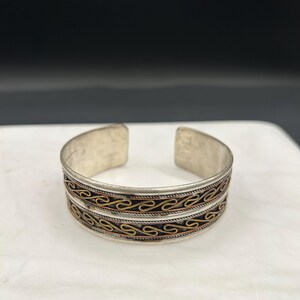 May include: Silver-tone cuff bracelet with decorative bands of gold and copper-colored filigree. The open-ended bracelet sits on a white surface with a dark background. The bracelet is a piece of jewelry.