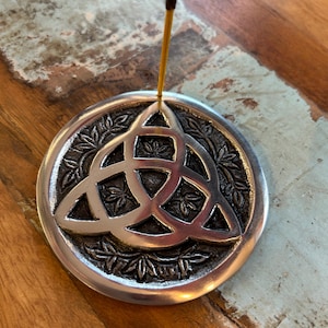 May include: A silver incense holder with a Celtic knot design. The round holder features a raised triquetra symbol in the centre, surrounded by a detailed leaf pattern. An incense stick is inserted into the holder.