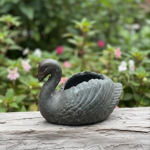 May include: A dark grey swan-shaped planter. The planter has a textured surface and a hollowed-out area for plants. The swan is perched on a weathered wooden surface, with a blurred background of green foliage and pink flowers.