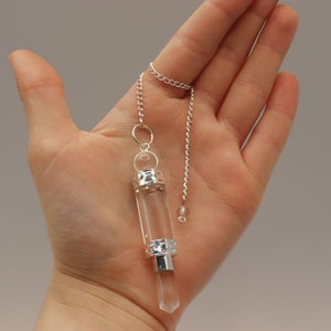 May include: A clear quartz crystal pendulum with a silver chain and two silver accents. The pendulum is hanging from a hand.