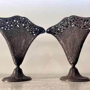 May include: Two dark grey metal decorative vases. Each vase features a flared top with an openwork design and an embossed pattern on the body. The vases stand on ornate, pedestal bases.