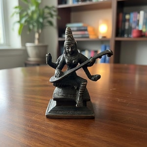 May include: A bronze-colored statue of a seated deity playing a stringed instrument. The figure has multiple arms and is adorned with detailed headwear and jewelry. The statue is on a square base and sits on a wooden table.