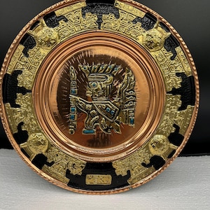 May include: A copper-toned decorative plate with a central figure holding a staff, surrounded by gold and black accents. The plate features intricate embossed designs and the word "PERU" at the bottom.