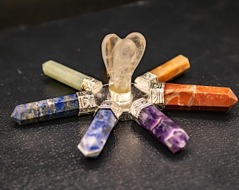 Könnte beinhalten: Ein Chakra-Set aus Kristallen mit sieben verschiedenen farbigen Steinen, die in einem Kreis um einen klaren Quarzengel angeordnet sind. Die Steine sind Amethyst, Lapislazuli, Sodalith, grüner Aventurin, Citrin, Karneol und roter Jaspis.
