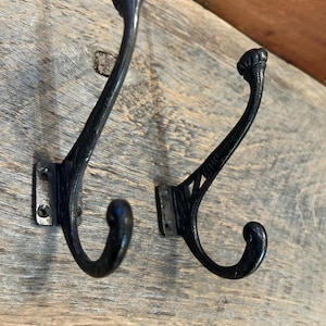 Może przedstawiać: Dwa czarne metalowe haczyki z ozdobnymi zwójami i kwadratową podstawą. Haczyki są zamontowane na drewnianej powierzchni.