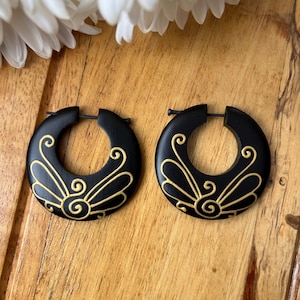 May include: A pair of black wooden hoop earrings with gold painted designs. The earrings are round and have a simple, elegant design.