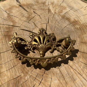 May include: Ornate antique brass drawer pull with a decorative floral design. The pull features a central fan-shaped element and curved arms with ring pulls. The hardware is set against a rustic wooden background.