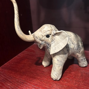 Può includere: Piccola statuetta di elefante fatta a mano con finitura bianco sporco testurizzata e sottili venature blu-grigie. L'elefante ha grandi orecchie, una lunga proboscide curva e piccole zanne. La statuetta è esposta su una superficie rossa.