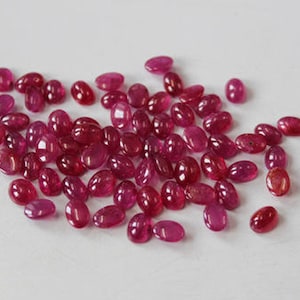 May include: A collection of oval-shaped, red gemstones. The gemstones are loose and scattered on a white surface.