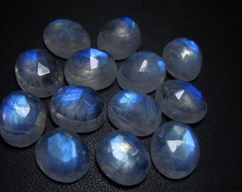 May include: A collection of faceted moonstone cabochons. The stones have a translucent, milky appearance with flashes of blue and white. The gemstones are oval-shaped and arranged on a dark surface, showcasing their natural beauty.