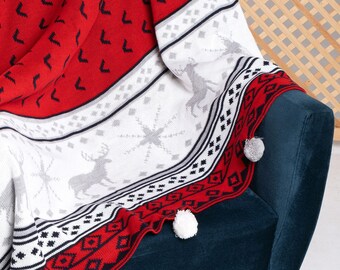 May include: Red and white knitted throw blanket with a reindeer pattern and white pom-poms. The blanket is draped over a dark blue armchair.