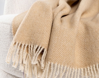 Könnte beinhalten: Eine weiche, beige Wolldecke mit Fischgrätenmuster und Fransenbesatz. Die Decke liegt über einem weißen Sessel.