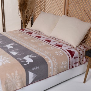 Pode incluir: Um cobertor bege e cinza com um padrão geométrico vermelho e branco com veados e flocos de neve. O cobertor está em uma cama com dois travesseiros brancos.