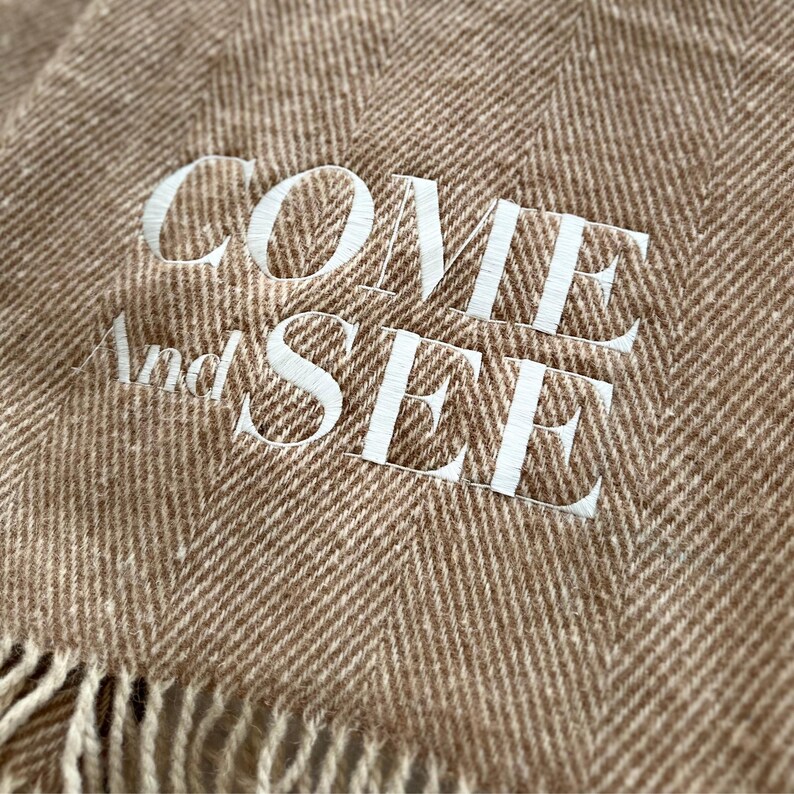 以下が含まれることがあります： 白で刺繍された「COME And SEE」の文字が入った、茶色と白のヘリンボーン柄の毛布です。