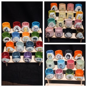 Könnte beinhalten: Eine Sammlung farbenfroher Keramikbecher, die jeweils mit einzigartigen Illustrationen von Anime-Charakteren verziert sind. Die Becher sind auf Holzregalen vor schwarzem Hintergrund angeordnet und zeigen eine Vielzahl von Designs und Farben.