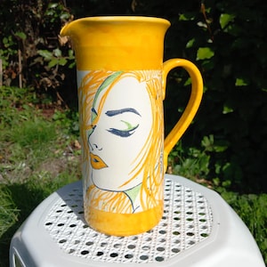 Peut inclure: Un pichet en céramique jaune avec un motif peint à la main d'un visage de femme aux yeux fermés et aux cheveux longs. Le pichet a une anse et une large ouverture.