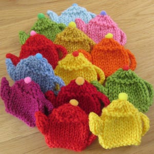 May include: A collection of 12 knitted teapot ornaments in various colours, including red, orange, yellow, green, blue, and purple. Each teapot has a small pom-pom on top.