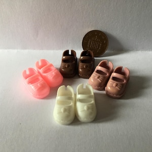 May include: A selection of miniature doll shoes in shades of pink, brown, and white. The shoes are displayed on a white surface, with a penny for scale. Styles include Mary Janes and sandals, perfect for a doll's wardrobe.