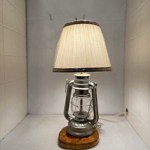 May include: A silver lantern lamp with a beige lampshade on a wooden base. The lamp is lit and the light shines through the glass.
