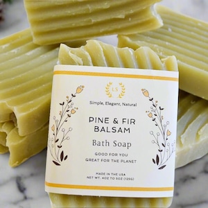May include: A stack of green bath soap bars with a white label that reads "Simple, Elegant, Natural" and "Pine & Fir Balsam Bath Soap" with a floral design. The label also says "Good for you, Great for the planet" and "Made in the USA".