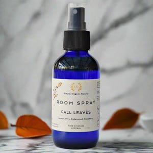 May include: A blue glass bottle with a black spray top and a white label that reads "ROOM SPRAY FALL LEAVES Lemon, Pine, Cedarwood, Rosemary MADE IN THE USA 4 oz/118ml".