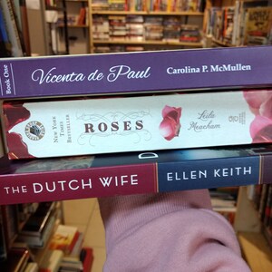 May include: Three stacked books with visible spines. The top book is purple with white text reading Vicenta de Paul by Carolina P. McMullen. The middle book is cream with red roses and text reading Roses by Leila Meacham. The bottom book is dark red and blue with white text reading The Dutch Wife by Ellen Keith.