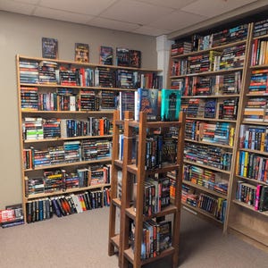 May include: A bookshelf filled with books, mostly paperbacks, in a variety of genres. The books are organized by color and size. There are also a few hardback books on the shelves. The bookshelf is in a room with a wooden floor and white walls.