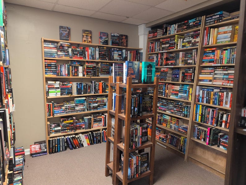 May include: A bookshelf filled with books, mostly paperbacks, in a variety of genres. The books are organized by color and size. There are also a few hardback books on the shelves. The bookshelf is in a room with a wooden floor and white walls.
